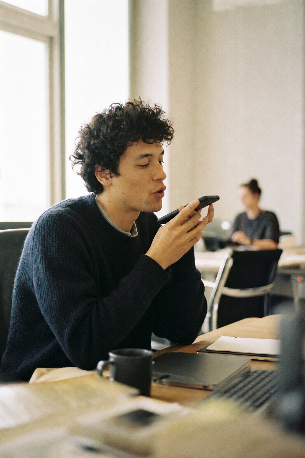 A person at a shared desk quietly mouthing into a phone in both hands.